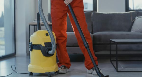 pexels-photo-6195273-6195273 A professional cleaner in red coveralls vacuums a contemporary living room with a yellow vacuum cleaner.
